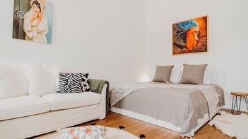 Un dormitorio moderno con un sofá blanco, una cama con manta color beige y dos cuadros en las paredes.