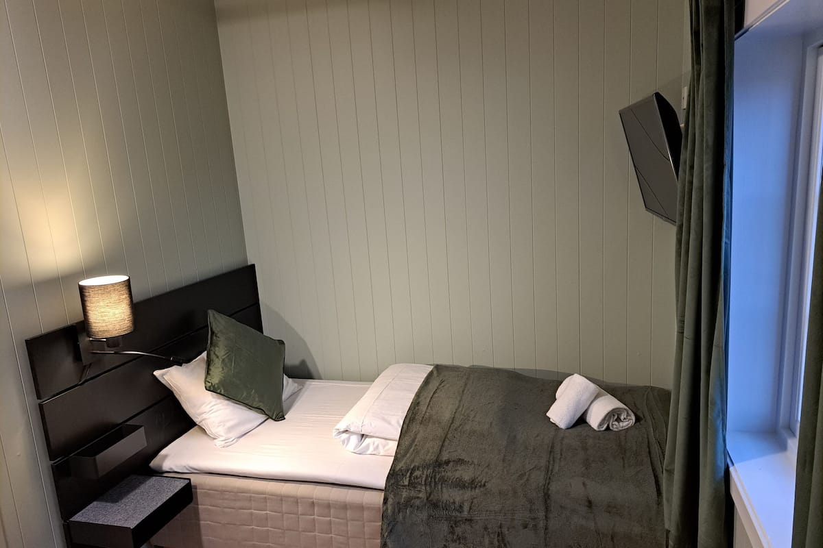 A small, neatly made bed with a green blanket, white pillows, and a bedside table with a lamp.