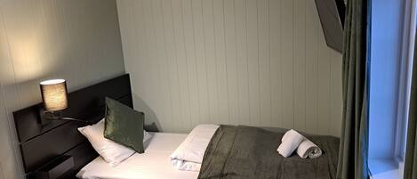 A small, neatly made bed with a green blanket, white pillows, and a bedside table with a lamp.