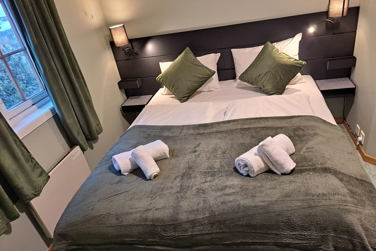 A neatly made bed with a dark green blanket, two rolled towels, and two green pillows.