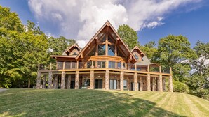 A large, multi-story log cabin with a balcony and stone pillars, surrounded by a grassy area and trees. - The Yellowstone | Elegance Meets Lakefront Luxury (Bracey)