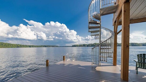 A wooden deck with a spiral staircase, a pool, and a view of a lake. - The Yellowstone | Elegance Meets Lakefront Luxury (Bracey)