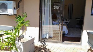 A room with a bed, a small table, and a chair. There is a window with curtains, and a plant is visible outside the window. - Nqabanqaba B&B  (Richards Bay)