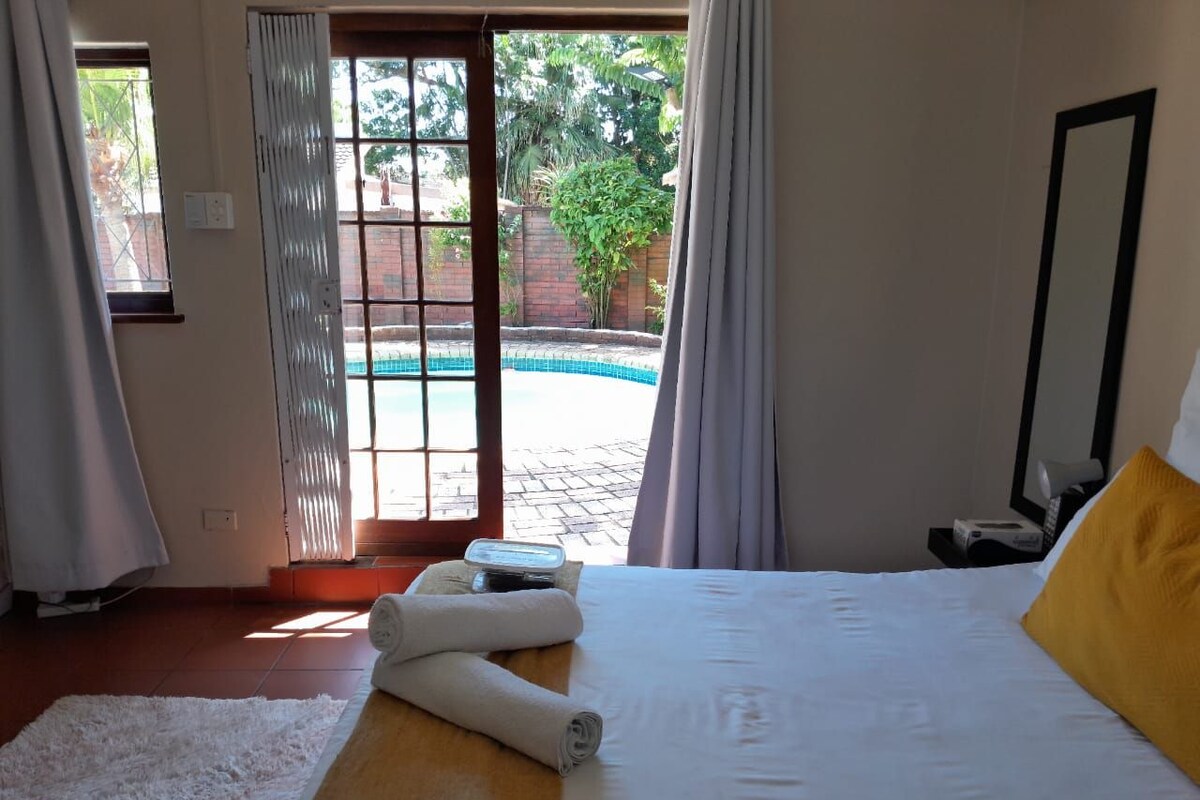 A bedroom with a bed, a white rug, a yellow pillow, and a view of a swimming pool.