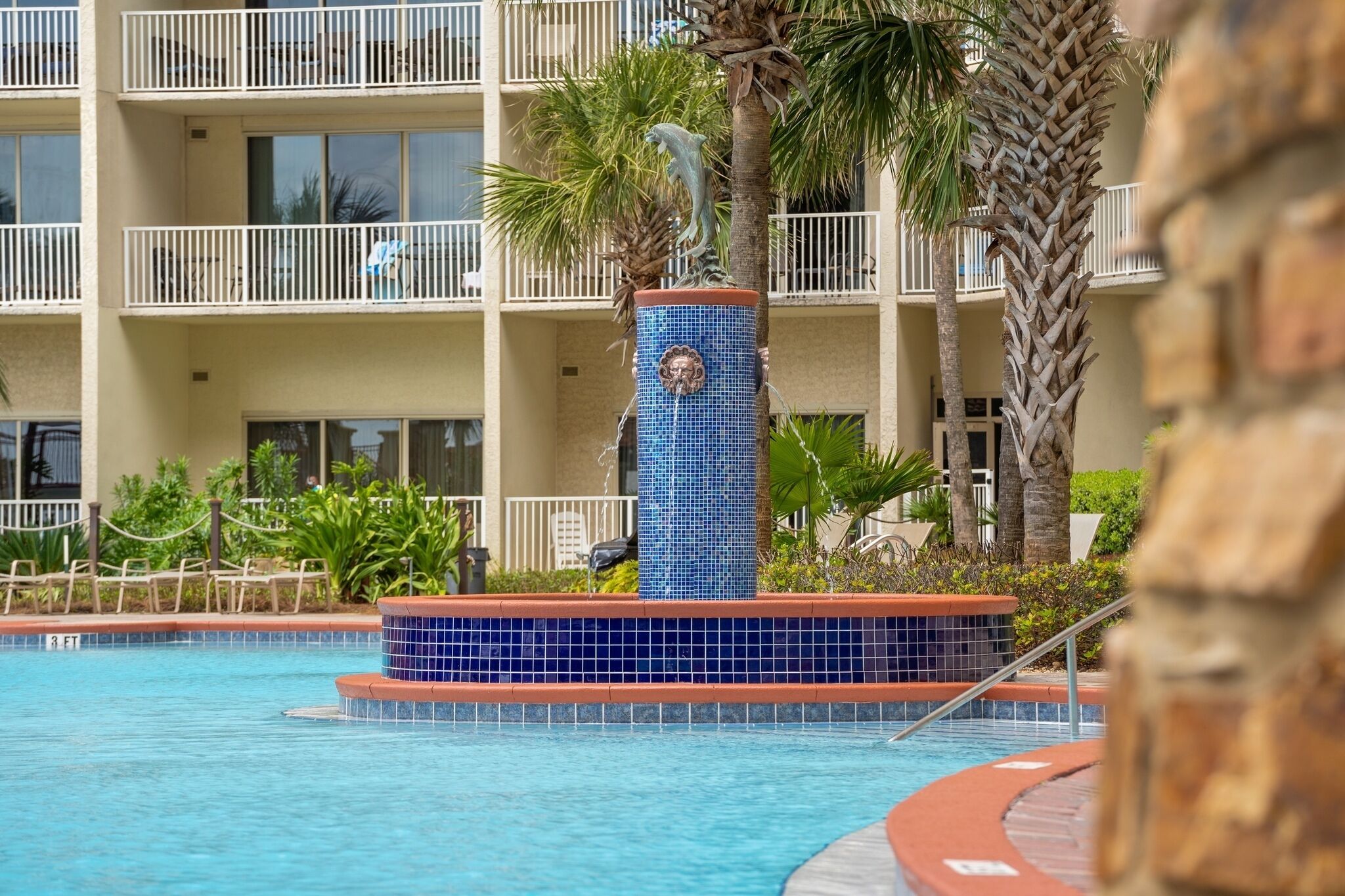 A hotel pool area with a decorative fountain, palm trees, and a building with balconies in the background.