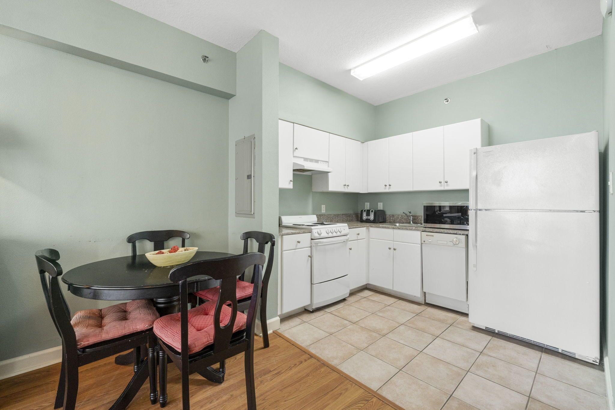 Une cuisine compacte avec des meubles blancs, une cuisinière et un réfrigérateur. On y trouve une table à manger avec des chaises et un saladier rempli de fruits.