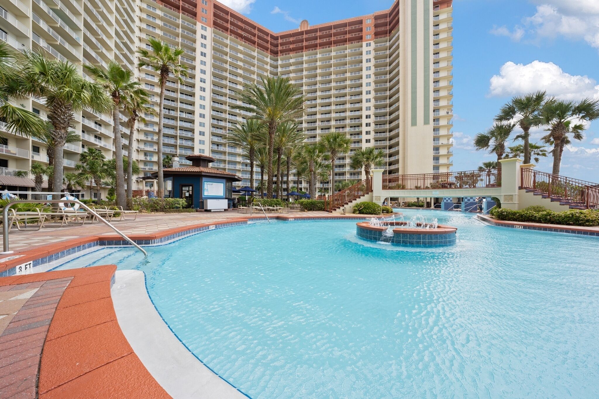 A swimming pool with a water fountain, surrounded by palm trees and a high-rise building in the background.