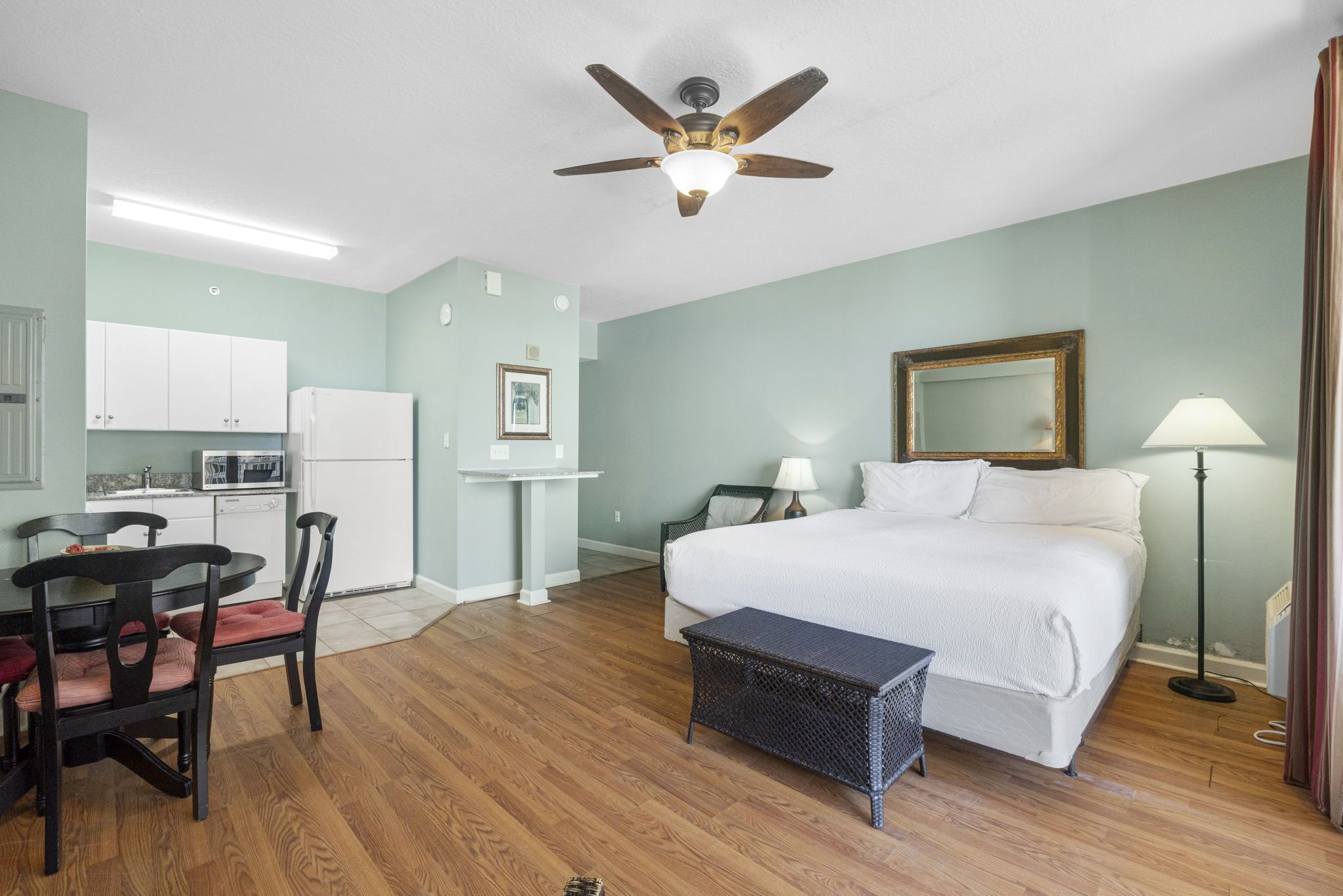 Une chambre d’hôtel équipée d’un lit, d’un bureau, d’une chaise, d’un ventilateur de plafond, d’une lampe et d’une kitchenette.