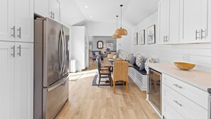 Una cocina moderna con armarios blancos, un refrigerador de acero inoxidable, una zona de comedor con mesa y sillas de madera, e iluminación colgante.