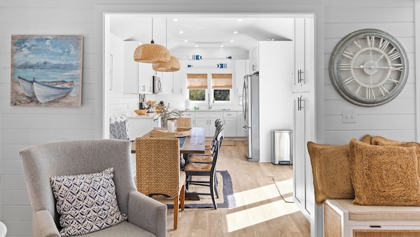 Una cocina moderna con armarios blancos, un comedor con mesa y sillas de madera, una silla de mimbre y un reloj de pared grande.