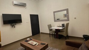 A living room with a flat-screen TV, a wooden coffee table, a brown sofa, a dining table with chairs, and a ceiling light fixture. - Guest House Quatre Saisons (Cotonou)