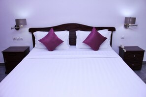Una cama bien tendida con ropa blanca y dos almohadas moradas, flanqueada por dos mesitas de noche con lámparas, en una habitación con paredes moradas.
