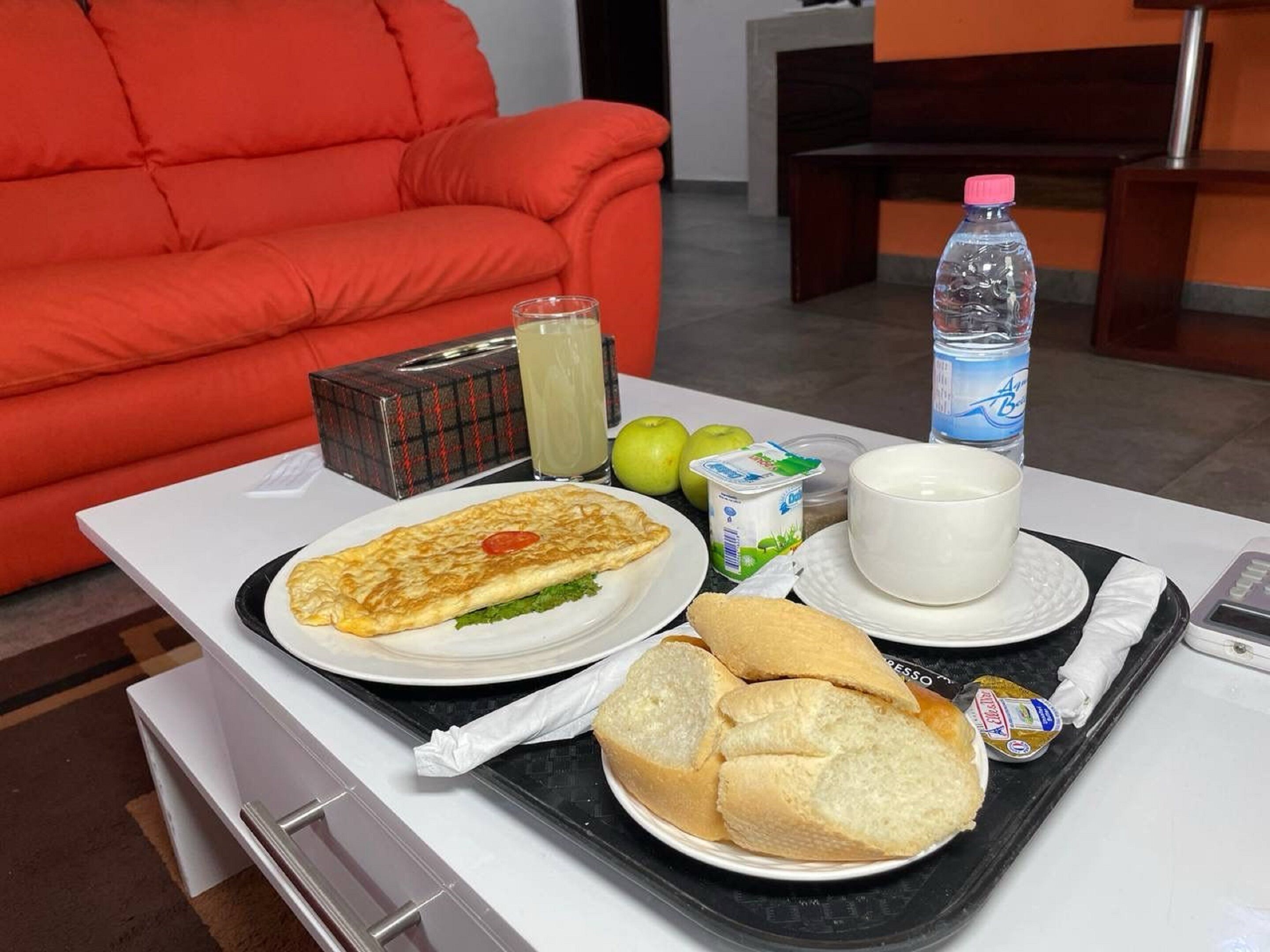 Bandeja de café da manhã com omelete, pão, maçãs, copo de suco, garrafa de água, pote de iogurte e um pequeno recipiente com manteiga.