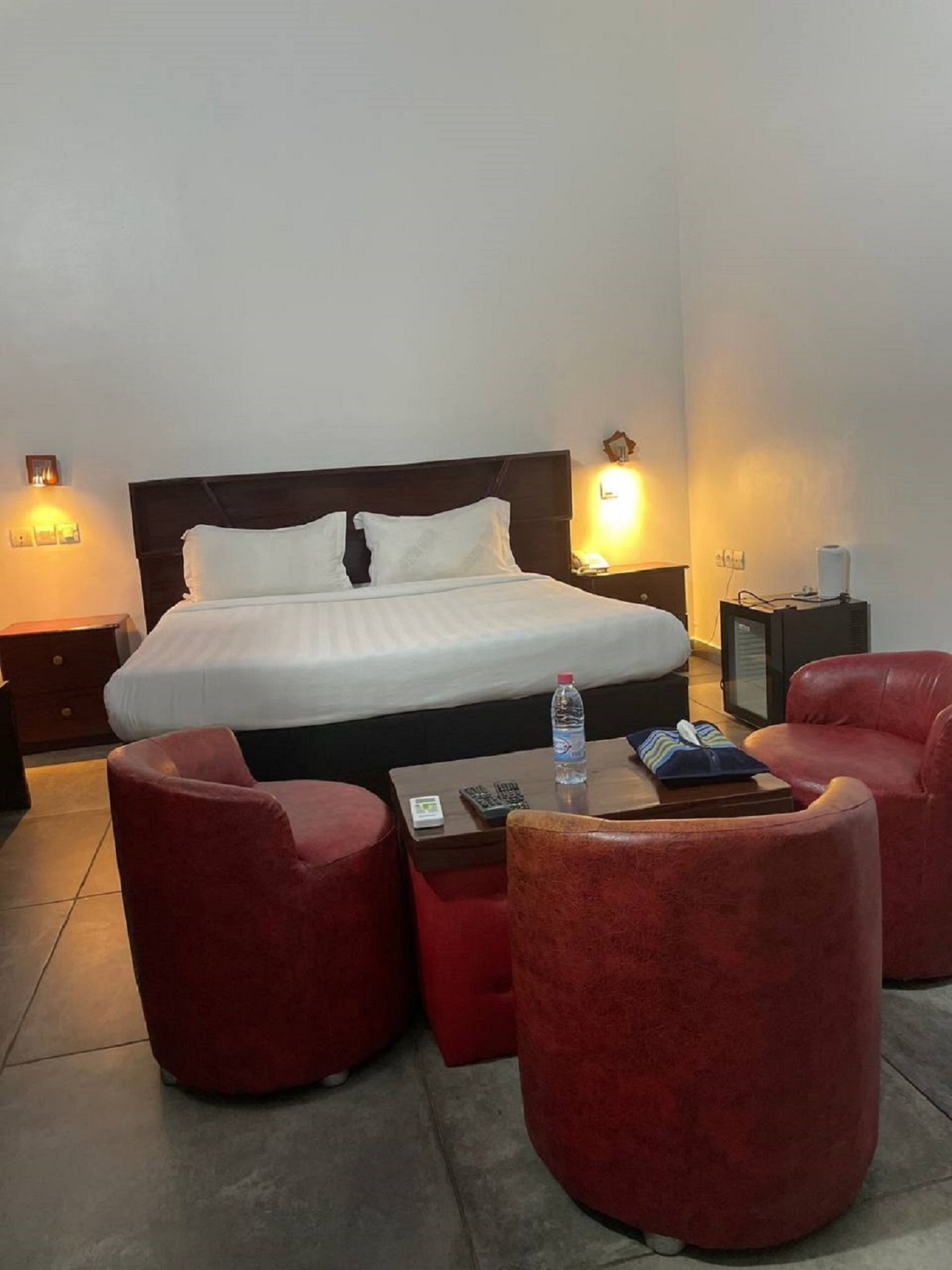Habitación de hotel con una cama grande, dos mesitas de noche con lámparas, un sillón rojo, una pequeña mesa con una botella y mandos a distancia, y un calentador fijado a la pared.