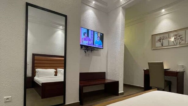 A hotel room with a large bed, a desk, a chair, a TV mounted on the wall, and a mirror. - Guest House Quatre Saisons (Cotonou)