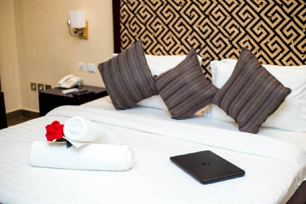 A neatly made bed with pillows, a rolled white towel, a red rose, and a tablet on the bed.