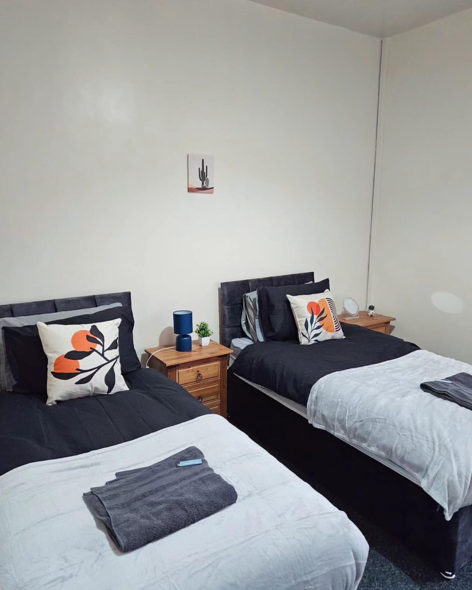 A hotel room with two beds, each with a black headboard, white bedding, and decorative pillows. There is a small table with a lamp and a plant between the beds.