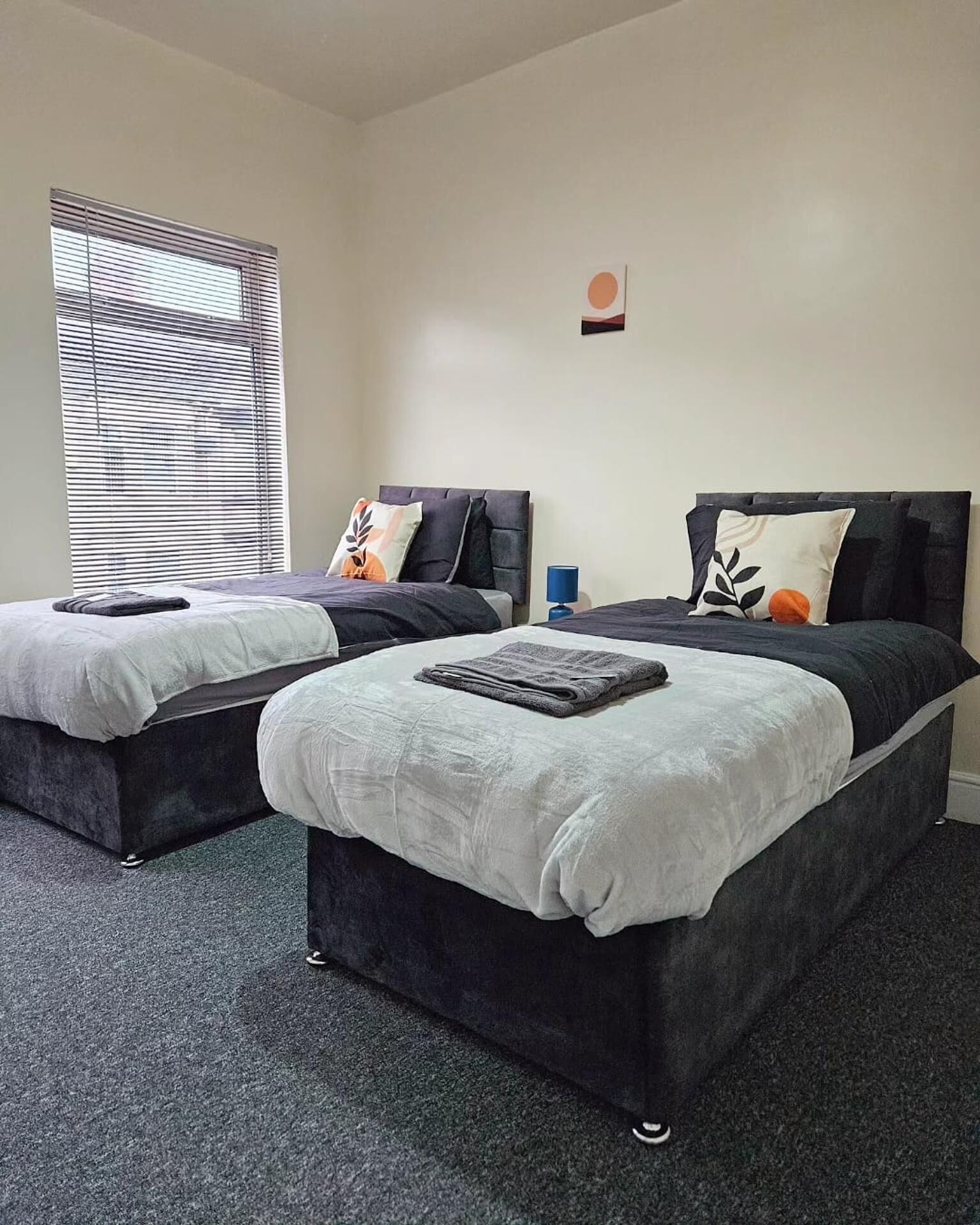 A hotel room with two beds, a large window with blinds, a small wall decoration, and a carpeted floor.