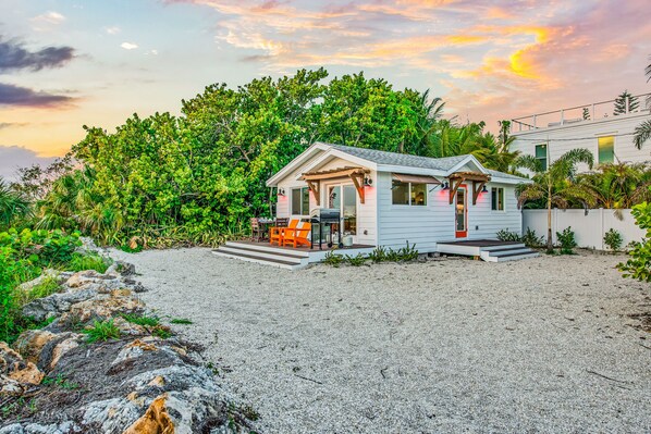 A small, single-story house with a porch, surrounded by lush greenery and a gravel driveway.