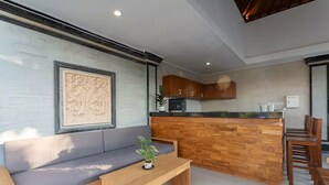 Een moderne woonkamer met een bank en een houten salontafel, een keuken met houten kasten en een bar, en een groot decoratief wandobject.