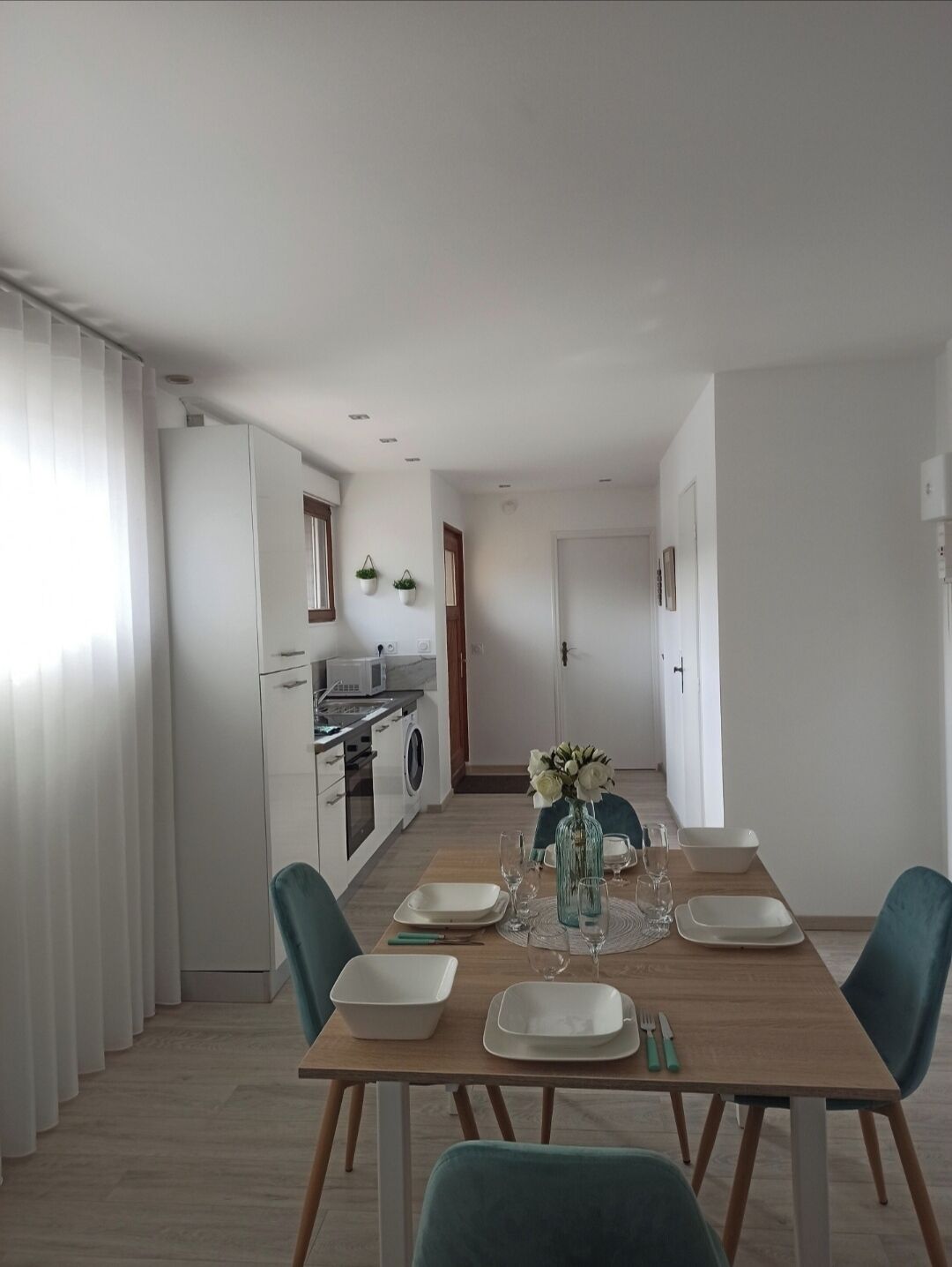 Un espace de repas moderne avec une table en bois dressée pour quatre personnes, une cuisine en arrière-plan et une vue sur le paysage urbain par la fenêtre.