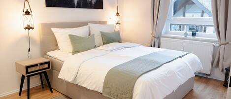 A bedroom with a bed, bedside tables, a window with curtains, a radiator, and a painting on the wall.