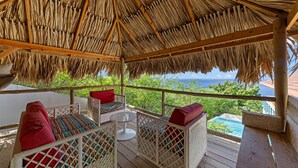 Un espacio al aire libre con techo, amueblado con sillas de mimbre y cojines rojos, con vistas a una piscina y al océano.