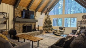 Een gezellige woonkamer met een stenen open haard, een grote kerstboom en uitzicht op het bos door grote ramen.