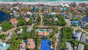 An aerial view of a residential area with houses, a tennis court, and a beach in the background.
