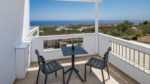A balcony with a table and chairs, overlooking a coastal landscape with buildings and a clear sky. - Hillside Elegance  (Santorini)