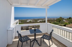 A balcony with a table and chairs, overlooking a coastal landscape with buildings and a clear sky.