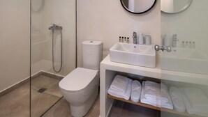 A modern bathroom with a glass-enclosed shower, a toilet, a sink with toiletries, and a shelf with folded towels. - Hillside Elegance  (Santorini)