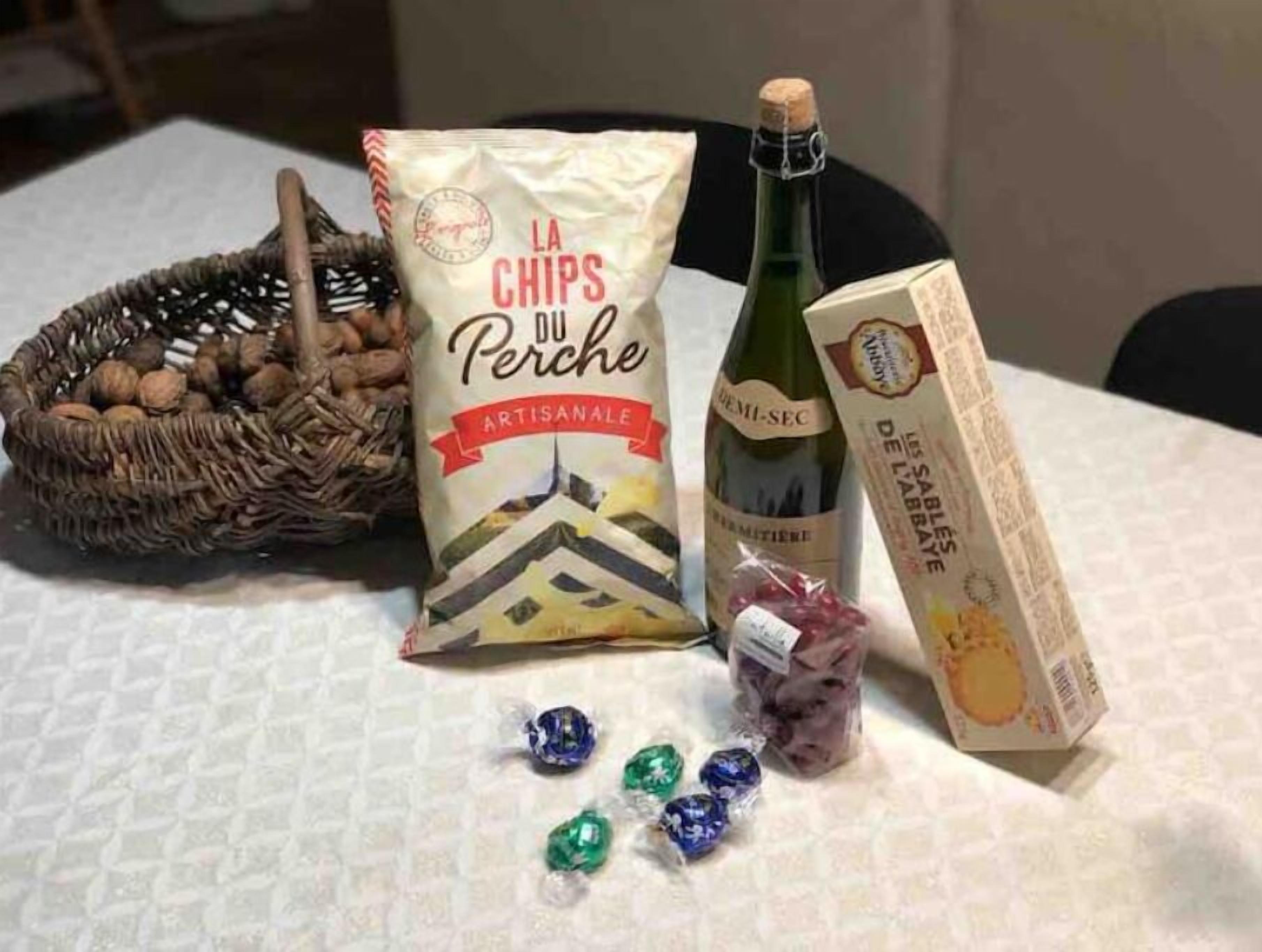Una mesa con una cesta tejida de nueces, una bolsa de papas artesanales, una botella de vino, una caja de bombones y una botella de agua con gas.