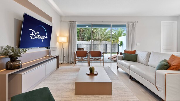 A modern living room with a flat-screen TV displaying the Disney+ logo, a white sofa, a coffee table with a cactus, and a balcony with outdoor furniture. - 5BR townhouse with pool near Disney area (Kissimmee)