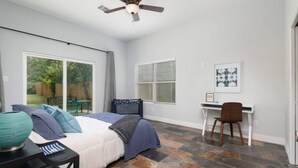 Un dormitorio con cama, escritorio y silla. Se ve una ventana con persianas y un ventilador de techo.
