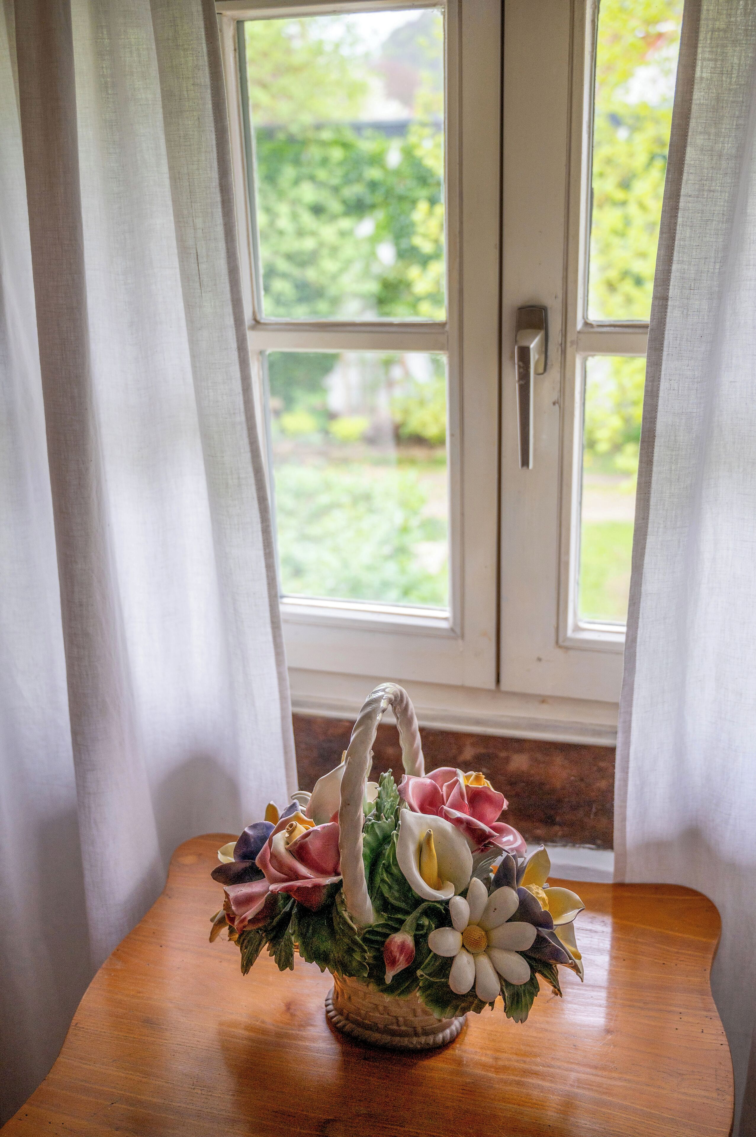 A wicker basket with a floral arrangement on a wooden table by a window with white curtains.