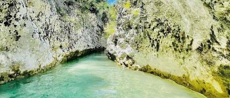 A narrow canyon with clear turquoise water, rocky walls, and sparse vegetation.