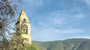 A stone church with a bell tower, surrounded by trees and a cobblestone path leading to a courtyard with a table and chairs.