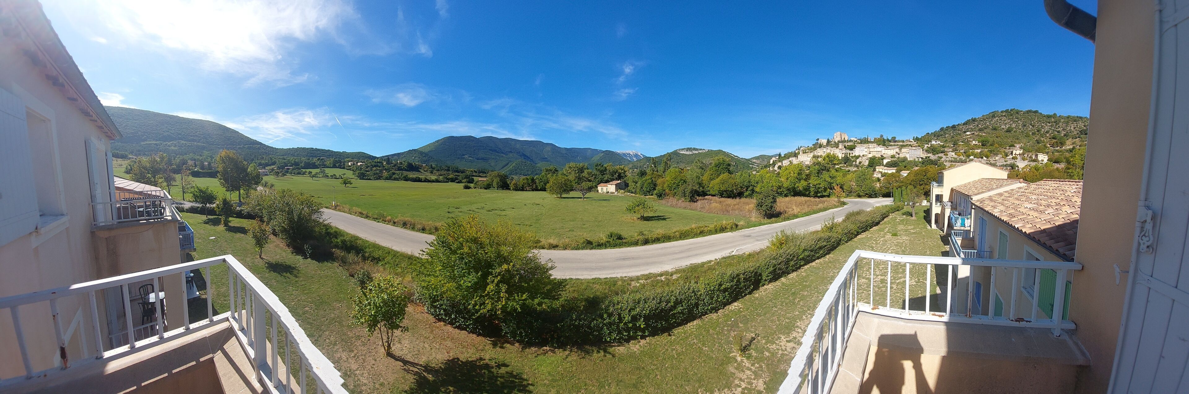 Un balcon offrant une vue sur une route sinueuse, des champs verdoyants et des montagnes lointaines.