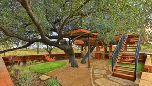 Une cabane dans les arbres avec un escalier en bois et une plateforme, entourée d’un jardin aménagé avec un sentier en pierre.