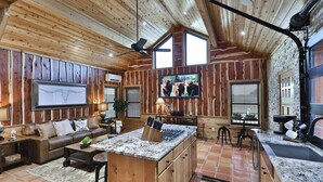 L’intérieur d’un chalet rustique est doté d’un plafond en bois, d’un grand téléviseur fixé au mur, d’un coin cuisine avec un îlot central et d’un espace salon confortable avec un canapé et une table basse.