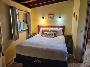Un lit bien fait, avec un coussin orné du mot «BONHEUR», une lampe de chevet et un tableau accroché au mur.
