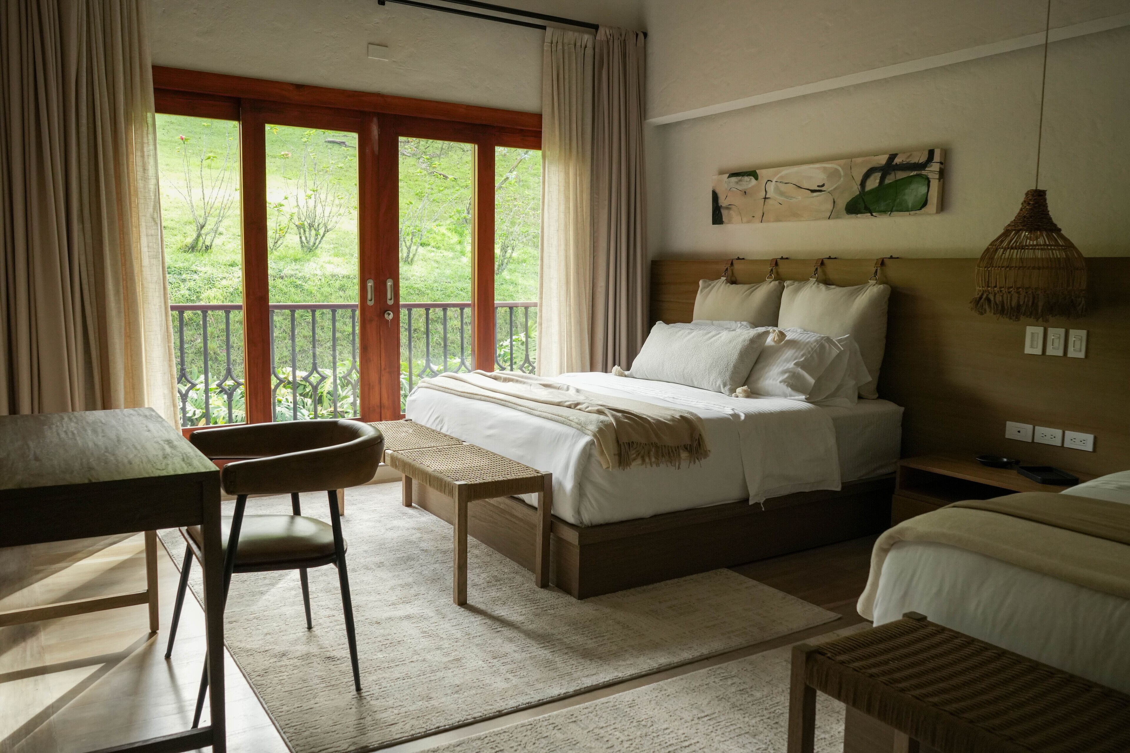 A bedroom with a bed, a desk, a chair, and a balcony with a view of greenery.