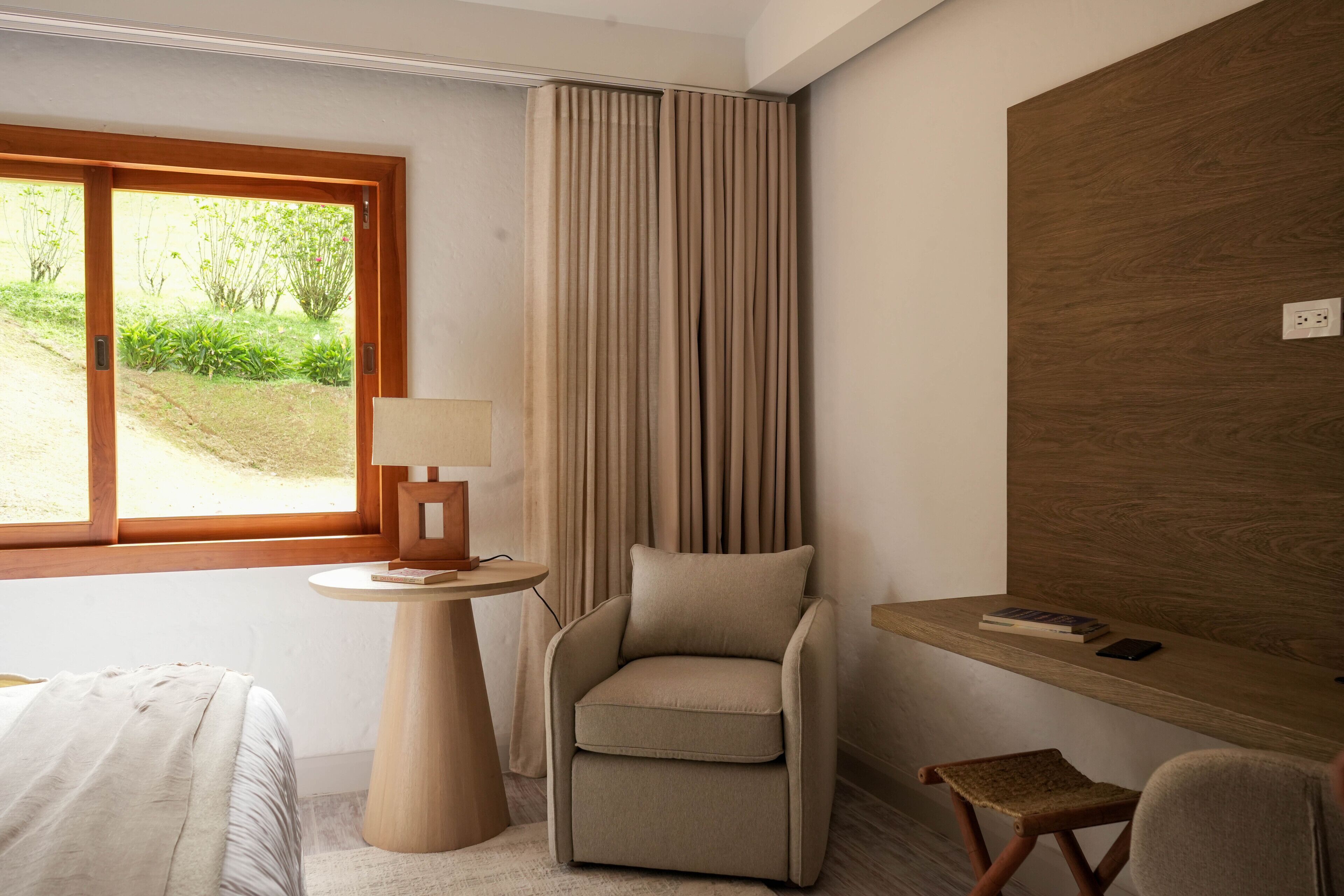 A hotel room with a bed, a chair, a small table with a lamp, a wooden desk, and a view of greenery outside.