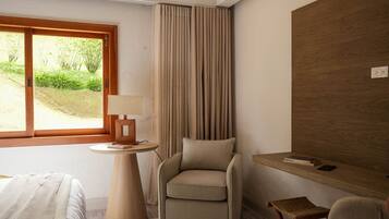 A hotel room with a bed, a chair, a small table with a lamp, a wooden desk, and a view of greenery outside.
