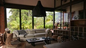 A cozy living room with a sofa, coffee table, and chairs, large windows with a view of lush greenery, and a bookshelf. - Mountains of Hope  (Rionegro)