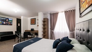 Une chambre d’hôtel équipée d’un lit, d’un bureau, d’une chaise, d’un canapé et d’un téléviseur diffusant une émission.