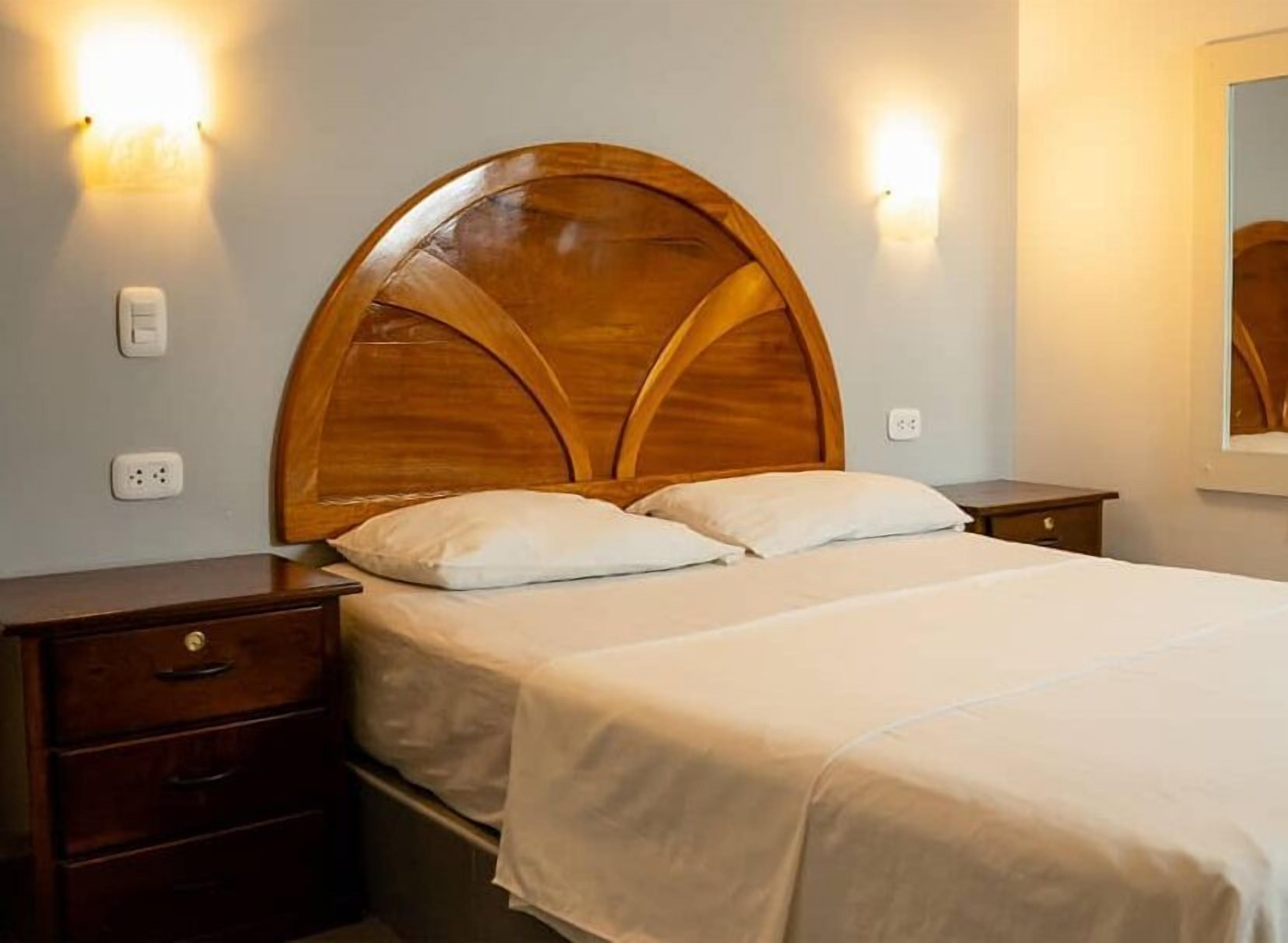 Una cama bien hecha con cabecera de madera, dos mesitas de noche e iluminación montada en la pared.