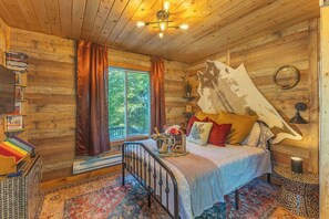 A cozy wooden cabin bedroom with a bed, a window with curtains, a bookshelf, and a unique wall art piece.