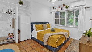 Ein Schlafzimmer mit Bett, Deckenventilator, Fenster mit Vorhängen, einem kleinen Tisch und einer Pflanze.